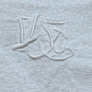 May include: Close-up of a white, textured fabric with an embroidered monogram. The monogram appears to be a stylized combination of letters, possibly initials, in a matching white thread. The fabric has a subtle, repeating pattern.