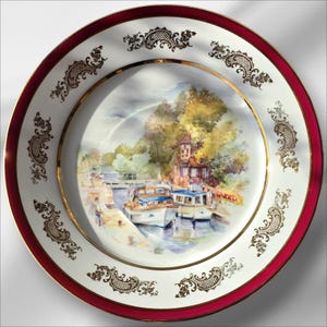 Może przedstawiać: Biały talerz porcelanowy z czerwonym rantem i złotą obwódką. Talerz przedstawia akwarelę przedstawiającą scenę kanału z łodziami, drzewami i domem.