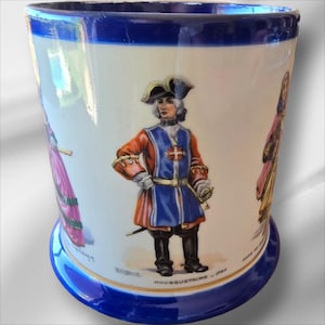 Puede incluir: Una taza de cerámica vintage con un borde y una base azules. La taza presenta una ilustración colorida de un hombre con un uniforme azul y rojo con una espada, y el texto "Mousquetaire - 1700".
