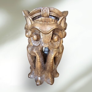 Könnte beinhalten: Eine Holzskulptur einer mythischen Kreatur mit aufwendigen Details. Die Skulptur ist in einem dunklen Braunton geschnitzt und hat eine strukturierte Oberfläche. Die Kreatur hat einen großen Kopf mit einer Krone und einen langen, fließenden Schwanz.