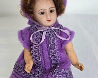 Può includere: Una piccola bambola di porcellana che indossa un vestito viola con bordi in pizzo e un cappello viola. La bambola ha i capelli castani e tratti dipinti.