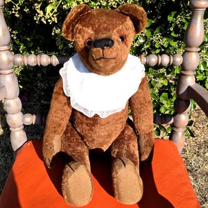 Peut inclure: Un ours en peluche marron avec des yeux sombres et un nez cousu noir, portant un col blanc bordé de dentelle. L'ours a une fourrure texturée et est assis en position verticale, montrant des coutures visibles sur ses pattes et ses membres.