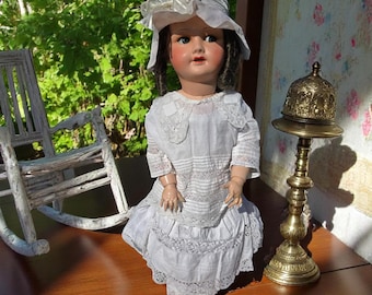 May include: A vintage porcelain doll wearing a white dress with lace trim and a matching hat. The doll has dark hair, blue eyes, and is posed on a wooden surface. A brass candle holder and a white rocking chair are in the background.