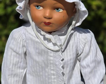 May include: A vintage doll with a painted face, blue eyes, and red lips, wearing a white bonnet and a white dress with lace trim. The doll's outfit includes a long-sleeved top and a skirt. The doll is a classic toy.