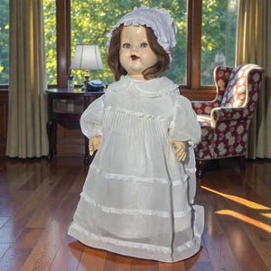 May include: A vintage doll with a porcelain face, brown hair, and blue eyes. The doll is dressed in a white dress with a ruffled collar and a matching bonnet. The doll is standing on a wooden floor.