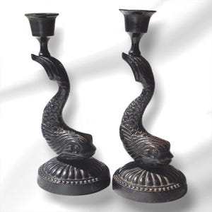 May include: Two dark bronze candle holders. Each has a dolphin design, with the body forming the stem and a circular base. The candle cups are at the top. The holders are approximately 20 cm tall.