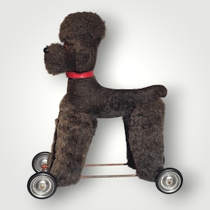 Può includere: Un giocattolo barboncino di peluche marrone con un collare rosso. Il giocattolo ha un corpo in tessuto strutturato e soffice pelliccia sulle zampe e sulla testa. Rotola su quattro piccole ruote con cerchi argentati e assi rossi. Il barboncino ha naso e occhi neri.