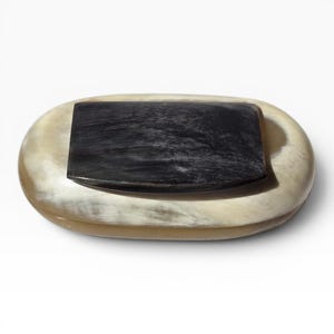 Puede incluir: Un objeto rectangular negro descansa sobre una bandeja ovalada de color crema. El objeto negro tiene una superficie texturizada. La bandeja tiene un aspecto liso y pulido, con variaciones de color.