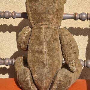 K&ouml;nnte beinhalten: Ein vintage brauner Teddyb&auml;r mit abgenutzter, pl&uuml;schiger Textur. Der B&auml;r sitzt auf einer roten Oberfl&auml;che.