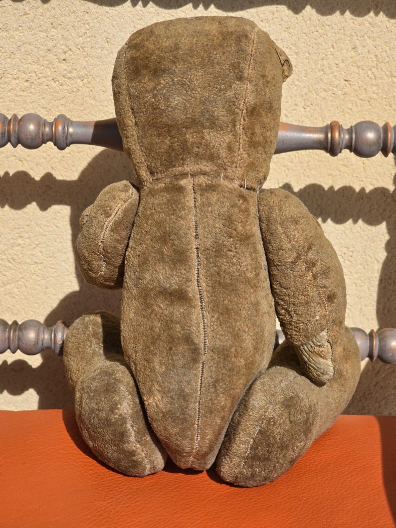 K&ouml;nnte beinhalten: Ein vintage brauner Teddyb&auml;r mit abgenutzter, pl&uuml;schiger Textur. Der B&auml;r sitzt auf einer roten Oberfl&auml;che.