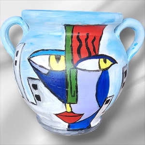 May include: A blue ceramic pot with two handles, painted with a stylized face in shades of blue, green, red, and yellow. The face has large eyes, a prominent nose, and a red mouth.