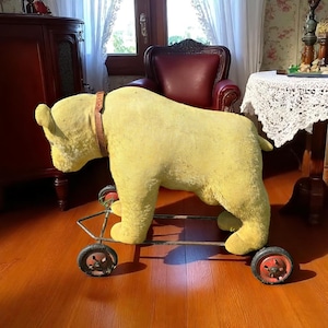 Puede incluir: Un juguete antiguo de peluche amarillo con forma de toro, con un collar de cuero marrón, montado sobre un marco de metal con cuatro ruedas. Las ruedas tienen detalles rojos. Un juguete clásico para montar para niños.