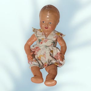 May include: A vintage baby doll with a painted face and a white romper with a colorful print. The doll has a glossy finish and is posed sitting down. The romper has ruffled sleeves and legs.