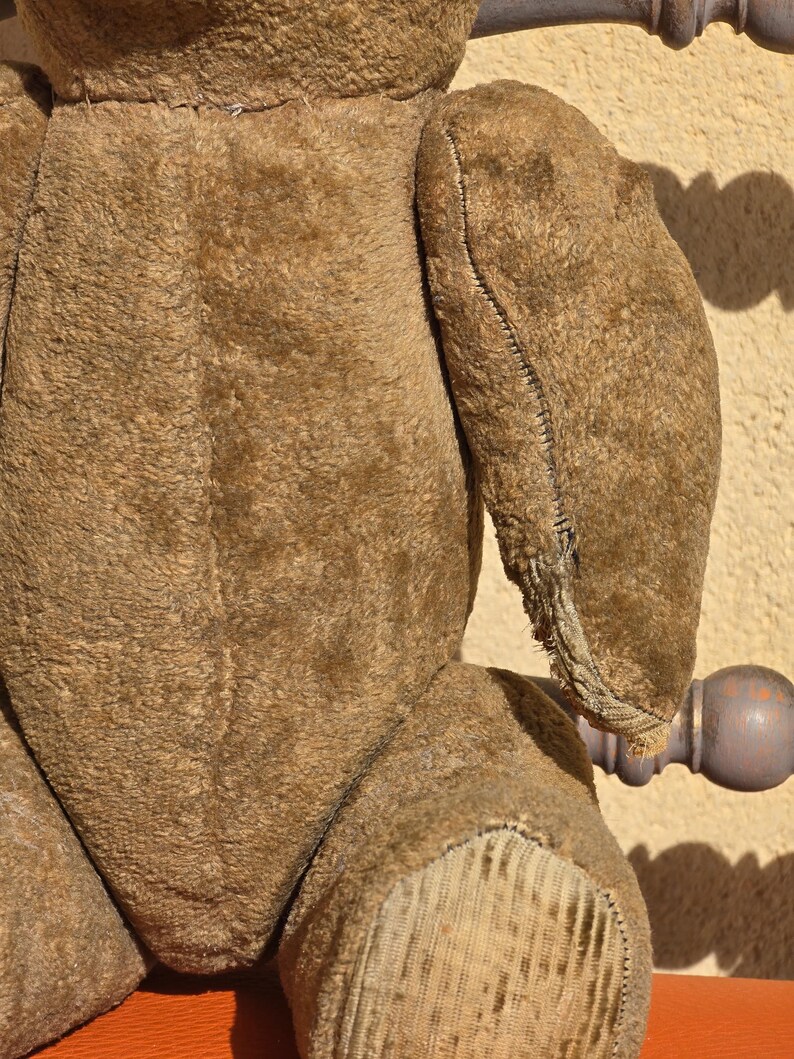 K&ouml;nnte beinhalten: Ein brauner Vintage-Teddyb&auml;r mit abgenutzter, flauschiger Textur. Der B&auml;r hat eine gen&auml;hte Naht in der Mitte des R&uuml;ckens und seine Arme sind angen&auml;ht. Die F&uuml;&szlig;e des B&auml;ren sind aus einem anderen Material gefertigt und haben ein abgenutztes, verblasstes Aussehen.