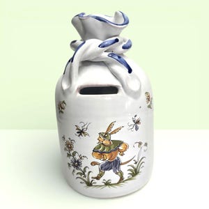 May include: White ceramic money bank shaped like a bag, with a slot for coins. The bank is decorated with a whimsical illustration of a person in a hat and floral and insect motifs. The top is tied with a blue-accented knot.