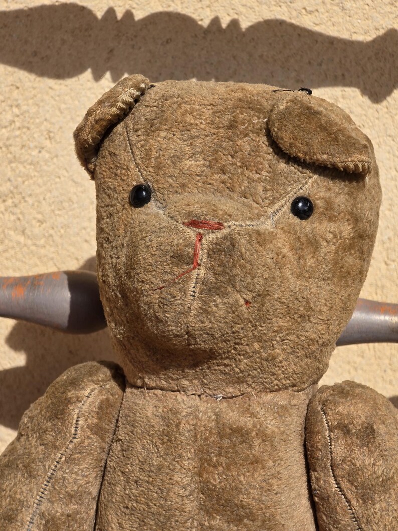 K&ouml;nnte beinhalten: Ein brauner Vintage-Teddyb&auml;r mit schwarzen Knopfaugen und einer roten gestickten Nase. Der B&auml;r sitzt auf einem Holzstuhl.