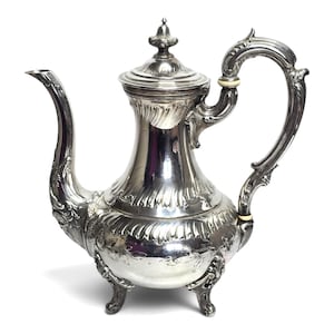 Puede incluir: Una cafetera antigua de color plateado con detalles ornamentados. La cafetera presenta un pico curvo, un asa grande y una tapa con un remate decorativo. El cuerpo está adornado con motivos florales y de volutas, y se apoya sobre tres pequeños pies.