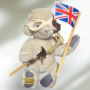 Op de afbeelding: Een lichtbeige teddybeer houdt een Union Jack-vlag en een pikhouweel vast. De beer draagt een masker en heeft grijze accenten op de poten. De vlag is blauw, rood en wit. Een gouden touw zit om de nek van de beer.