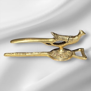 May include: A gold-coloured nutcracker with a bird-shaped design. The nutcracker features a lever and a base with floral detailing. The bird's beak acts as the cracking mechanism. The overall design is ornate and decorative.