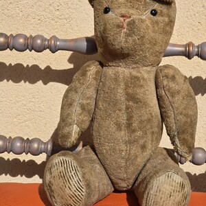 K&ouml;nnte beinhalten: Ein vintage brauner Teddyb&auml;r mit schwarzen Knopfaugen, der auf einem roten Stuhl sitzt. Der B&auml;r hat ein abgenutztes, verblasstes Aussehen und ist aus Pl&uuml;schstoff.