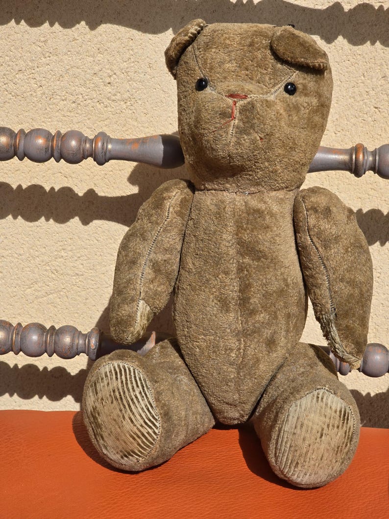 K&ouml;nnte beinhalten: Ein vintage brauner Teddyb&auml;r mit schwarzen Knopfaugen, der auf einem roten Stuhl sitzt. Der B&auml;r hat ein abgenutztes, verblasstes Aussehen und ist aus Pl&uuml;schstoff.