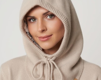 May include: A beige knit hood with a drawstring closure and a small leather tag. The hood is made of a soft, textured material and has a ribbed knit trim around the face. The interior lining has a floral pattern.