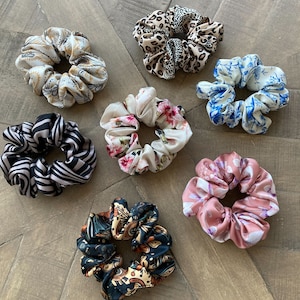 Könnte beinhalten: Ein Set aus acht bunten Haargummis mit verschiedenen Mustern, darunter Blumen-, Tier- und Paisley-Designs. Die Haargummis sind aus weichem Stoff gefertigt und perfekt, um jeder Frisur einen Hauch von Stil zu verleihen.