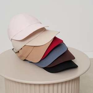 Op de afbeelding: Een stapel baseballpetten in verschillende kleuren, waaronder lichtroze, beige, rood, blauw, bruin en zwart. De petten worden gepresenteerd op een ronde, lichtgekleurde tafel, die hun verschillende tinten en stijlen benadrukt.