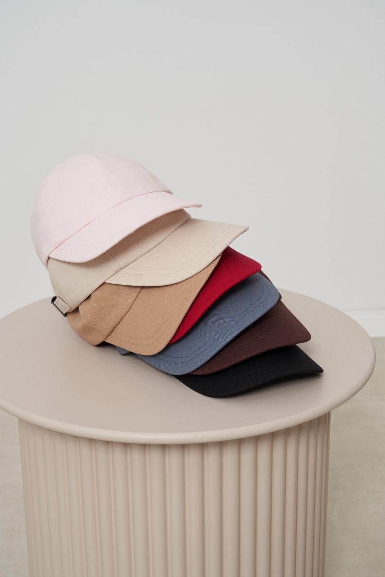 Op de afbeelding: Een stapel baseballpetten in verschillende kleuren, waaronder lichtroze, beige, rood, blauw, bruin en zwart. De petten worden gepresenteerd op een ronde, lichtgekleurde tafel, die hun verschillende tinten en stijlen benadrukt.
