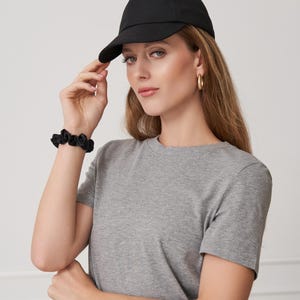 Op de afbeelding: Een zwarte baseballpet met een gebogen klep. De pet wordt gedragen met een grijs t-shirt en een zwarte scrunchie. Gouden oorringen maken de look compleet. De outfit is eenvoudig en casual.