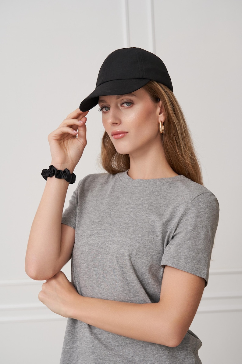 Op de afbeelding: Een zwarte baseballpet met een gebogen klep. De pet wordt gedragen met een grijs t-shirt en een zwarte scrunchie. Gouden oorringen maken de look compleet. De outfit is eenvoudig en casual.