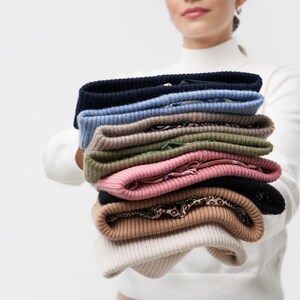 Puede incluir: Una pila de ocho su&eacute;teres de punto en varios colores, incluyendo beige, marr&oacute;n, rosa, verde, azul y negro. Los su&eacute;teres est&aacute;n doblados cuidadosamente y est&aacute;n siendo sostenidos por una persona.