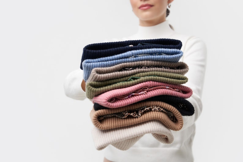 Puede incluir: Una pila de ocho su&eacute;teres de punto en varios colores, incluyendo beige, marr&oacute;n, rosa, verde, azul y negro. Los su&eacute;teres est&aacute;n doblados cuidadosamente y est&aacute;n siendo sostenidos por una persona.