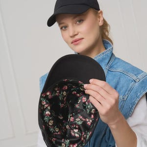 Op de afbeelding: Een zwarte baseballpet met een gebogen klep. De binnenvoering heeft een bloemenpatroon met rode, roze en groene bloemen op een zwarte achtergrond. De pet is gemaakt van een glad, effen materiaal.