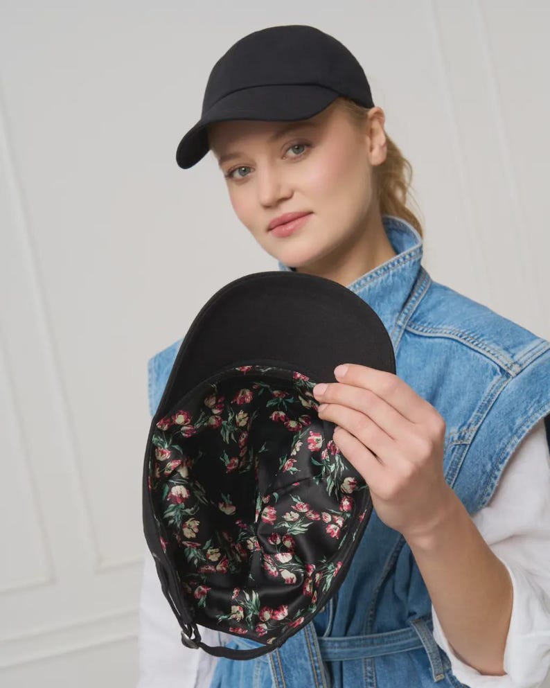 Op de afbeelding: Een zwarte baseballpet met een gebogen klep. De binnenvoering heeft een bloemenpatroon met rode, roze en groene bloemen op een zwarte achtergrond. De pet is gemaakt van een glad, effen materiaal.