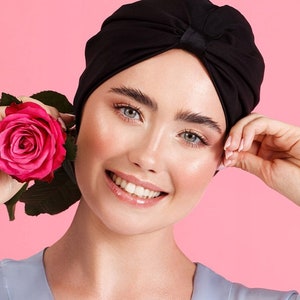 May include: A woman with a black turban is smiling and holding a pink rose. She is wearing a light blue shirt and has a tattoo on her arm.