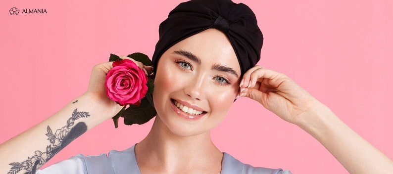 May include: A woman with a black turban is smiling and holding a pink rose. She is wearing a light blue shirt and has a tattoo on her arm.