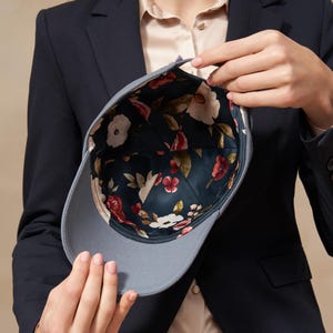 Op de afbeelding: Een grijze baseballcap met een bloemenpatroon aan de binnenkant. De cap wordt opengehouden en toont een donkergroene stof met witte en rode bloemen. De buitenkant van de cap is effen grijs.