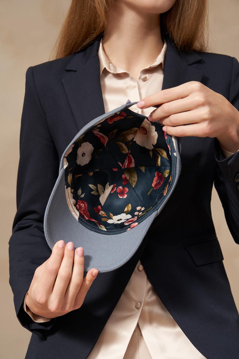 Op de afbeelding: Een grijze baseballcap met een bloemenpatroon aan de binnenkant. De cap wordt opengehouden en toont een donkergroene stof met witte en rode bloemen. De buitenkant van de cap is effen grijs.