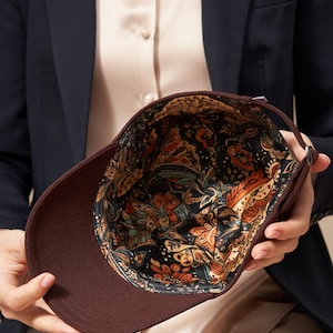 Op de afbeelding: Een bruine baseballcap met een patroon aan de binnenkant. De buitenkant van de cap is effen donkerbruin, terwijl de binnenkant een kleurrijk bloemmotief heeft met oranje, blauwe en gele accenten op een zwarte achtergrond. De cap wordt opengehouden.