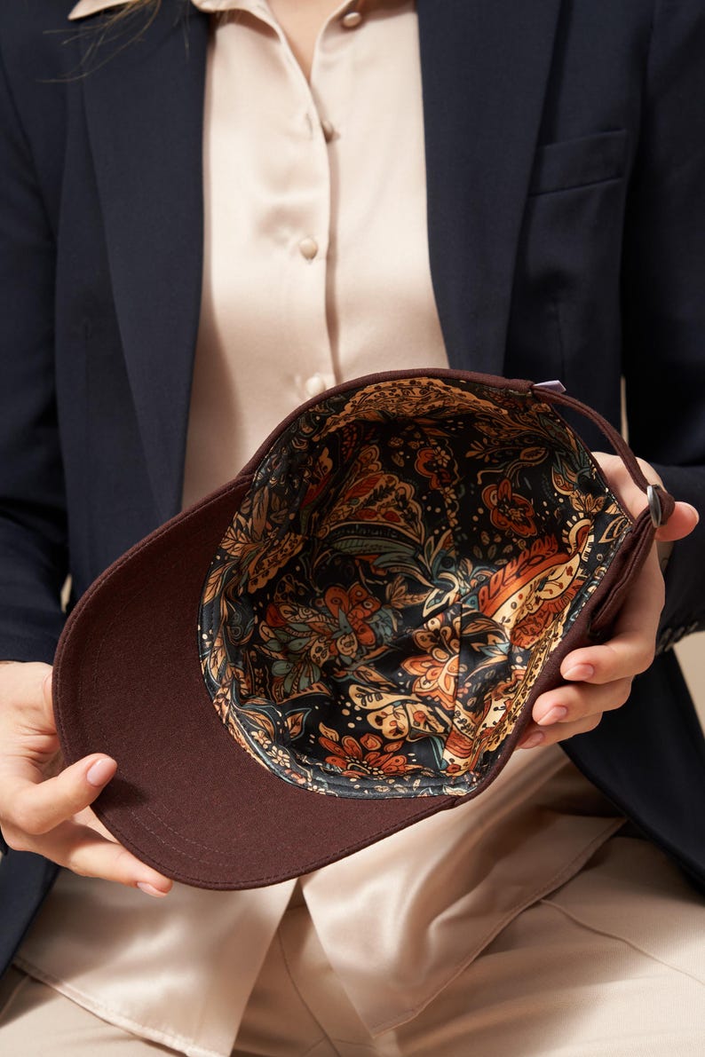 Op de afbeelding: Een bruine baseballcap met een patroon aan de binnenkant. De buitenkant van de cap is effen donkerbruin, terwijl de binnenkant een kleurrijk bloemmotief heeft met oranje, blauwe en gele accenten op een zwarte achtergrond. De cap wordt opengehouden.