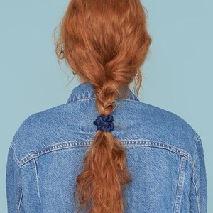 以下が含まれることがあります： 青いデニムジャケットを着た、長く、巻き毛の赤い髪の人の写真。髪は、ネイビーブルーのシュシュ2つでポニーテールにまとめられています。