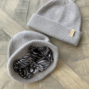 Puede incluir: Dos gorros de punto grises. Un gorro tiene un forro de estampado de cebra en blanco y negro. El gorro tiene una etiqueta de cuero marr&oacute;n con la palabra "ALMANIA" en ella.