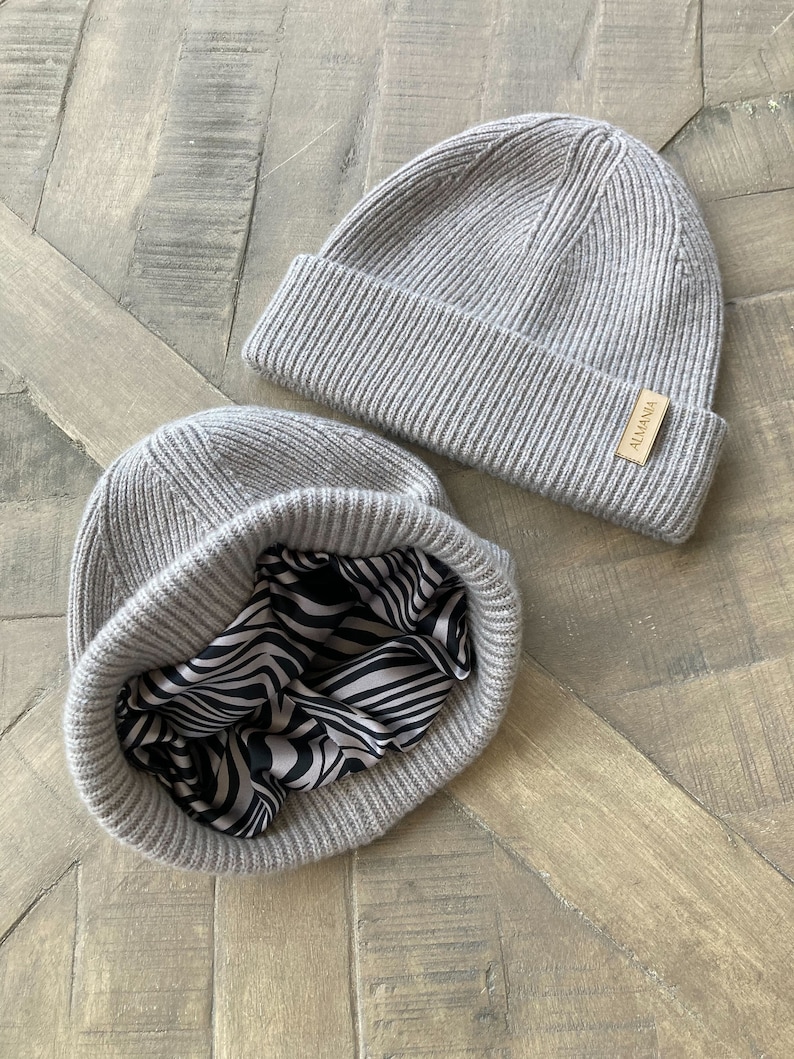 Puede incluir: Dos gorros de punto grises. Un gorro tiene un forro de estampado de cebra en blanco y negro. El gorro tiene una etiqueta de cuero marr&oacute;n con la palabra "ALMANIA" en ella.