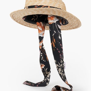 Puede incluir: Un sombrero de paja de ala ancha con una cinta floral negra y naranja atada alrededor de la corona. La cinta es larga y fluye por la parte posterior del sombrero.