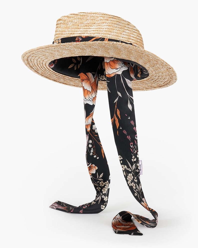 Puede incluir: Un sombrero de paja de ala ancha con una cinta floral negra y naranja atada alrededor de la corona. La cinta es larga y fluye por la parte posterior del sombrero.
