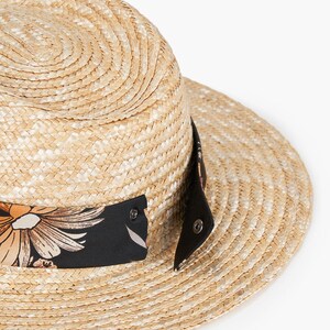 Puede incluir: Un sombrero de paja con una banda negra y un estampado floral. El sombrero tiene un ala ancha y una corona plana.