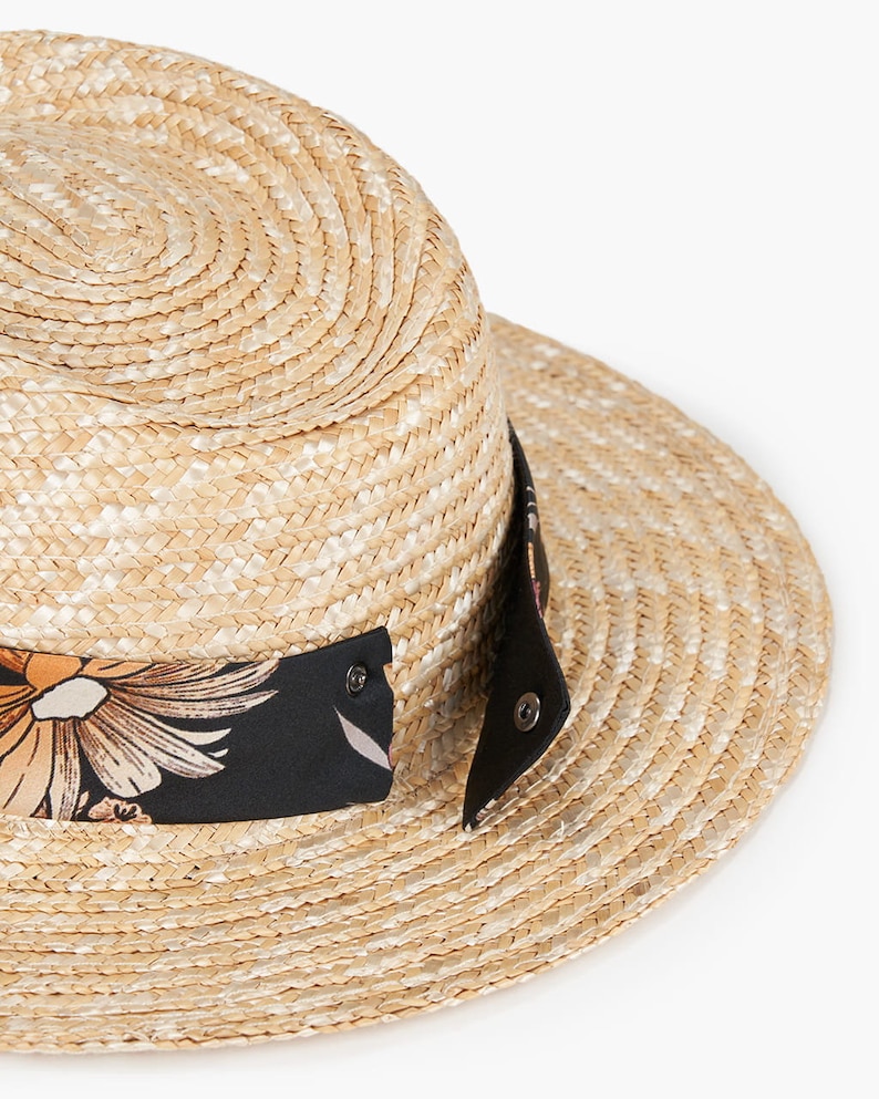Puede incluir: Un sombrero de paja con una banda negra y un estampado floral. El sombrero tiene un ala ancha y una corona plana.