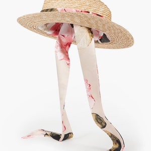 Puede incluir: Un sombrero de paja de ala ancha con una cinta floral atada alrededor de la corona. La cinta es larga y fluye por la parte posterior del sombrero.