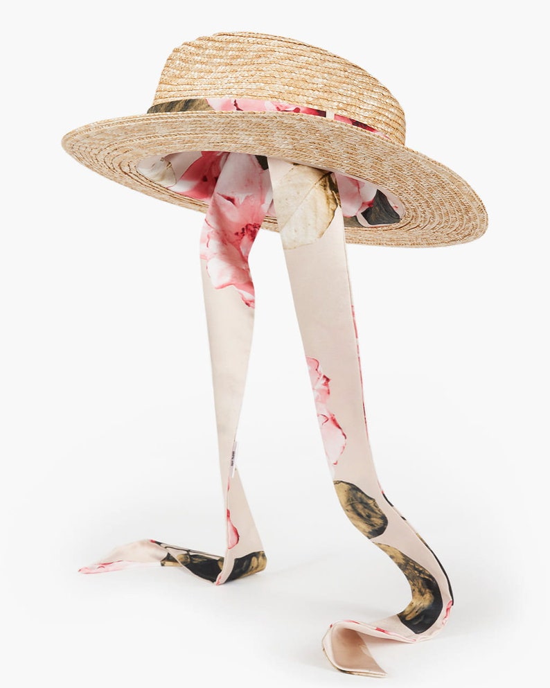 Puede incluir: Un sombrero de paja de ala ancha con una cinta floral atada alrededor de la corona. La cinta es larga y fluye por la parte posterior del sombrero.
